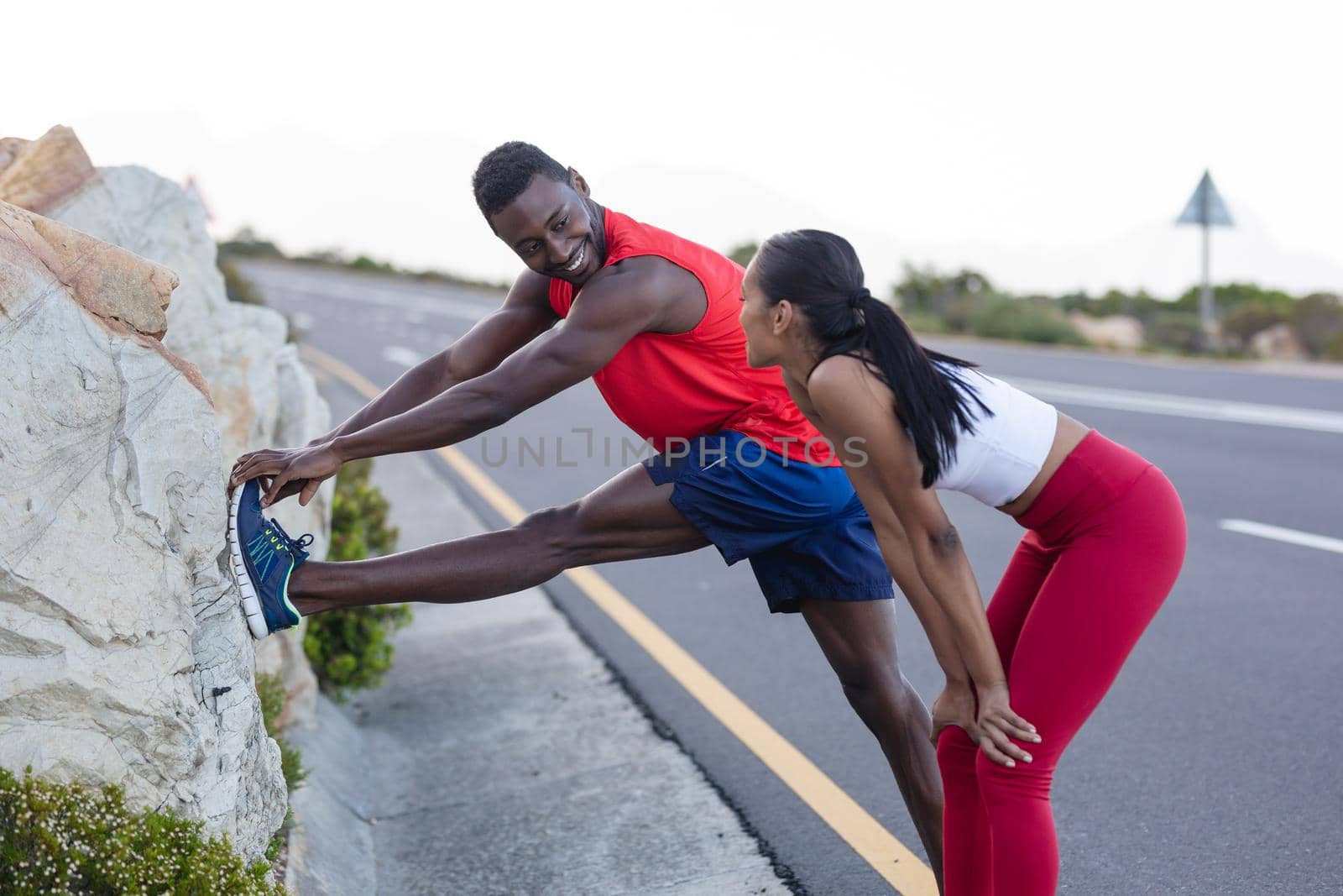 Fit african american man in sportswear stretching while woman is standing on a coastal road. healthy lifestyle, exercising in nature.