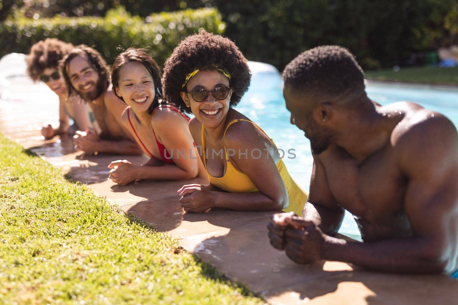 Diverse group of friends smiling and leaning on the poolside. Hanging out and relaxing outdoors in summer.