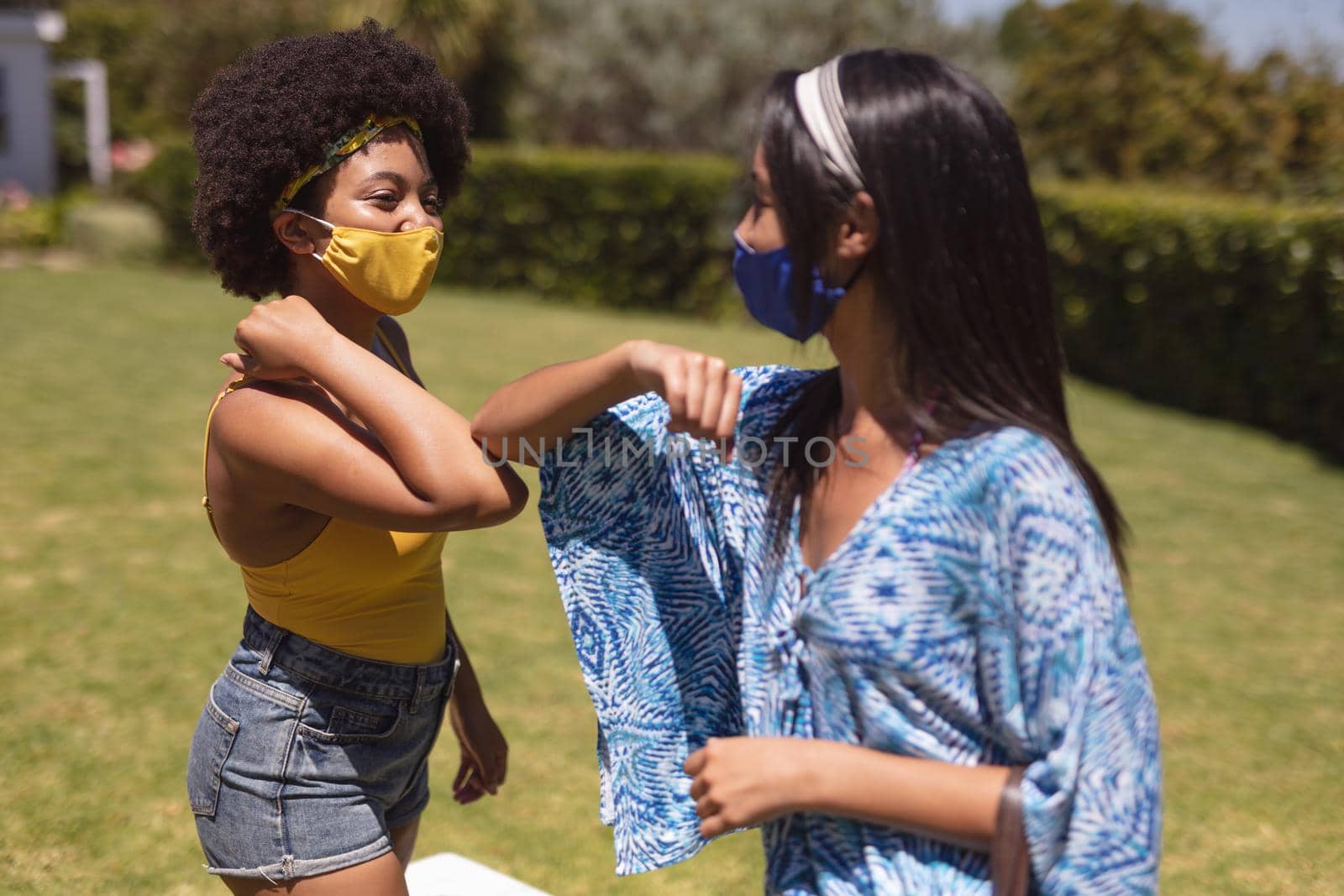 Two diverse female friends wearing face masks bumping elbows at a pool party. Health and hygiene precautions while hanging out and relaxing outdoors in summer during coronavirus covid 19 pandemic.