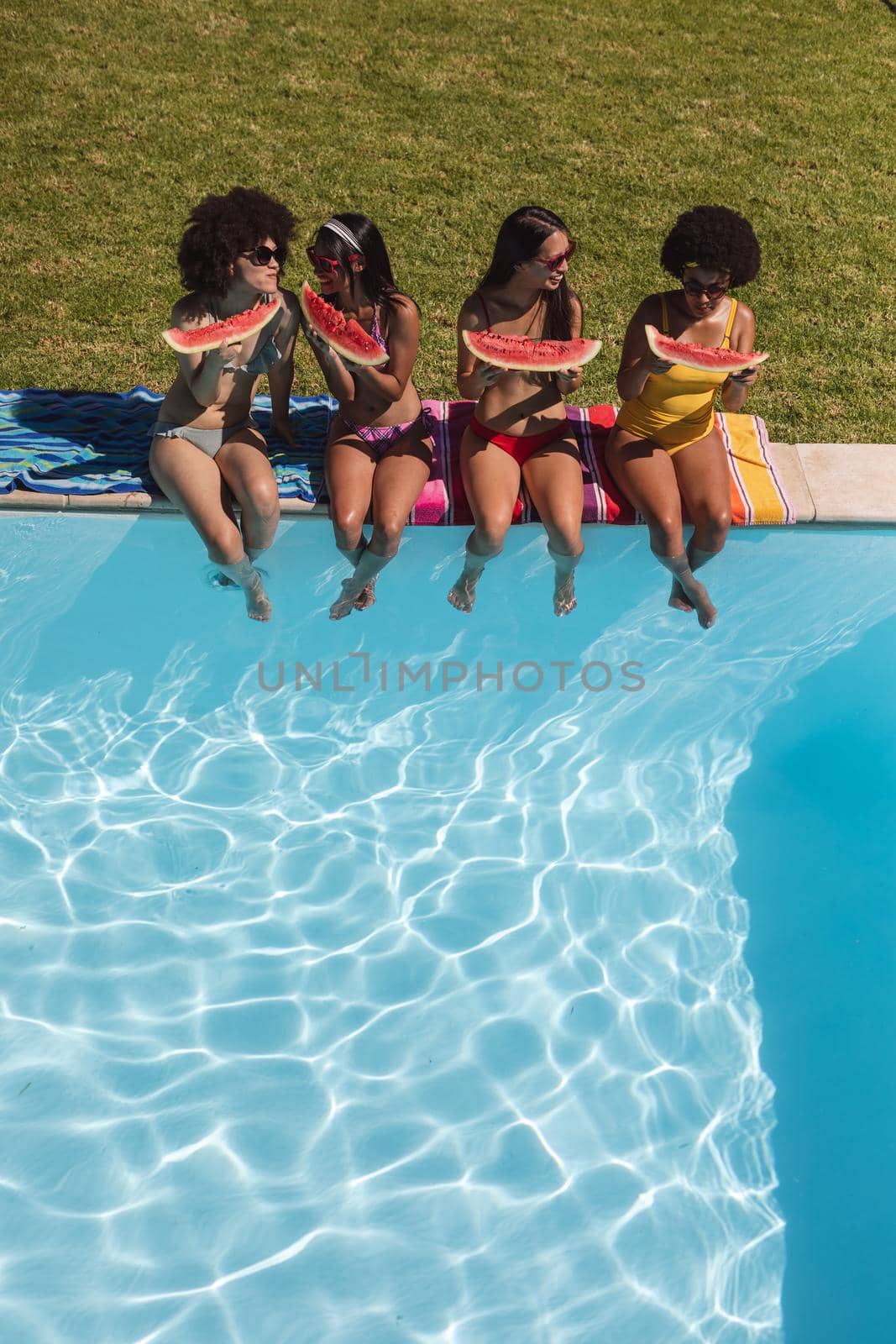 Diverse group of female friends eating watermelon sitting at the poolside talking by Wavebreakmedia