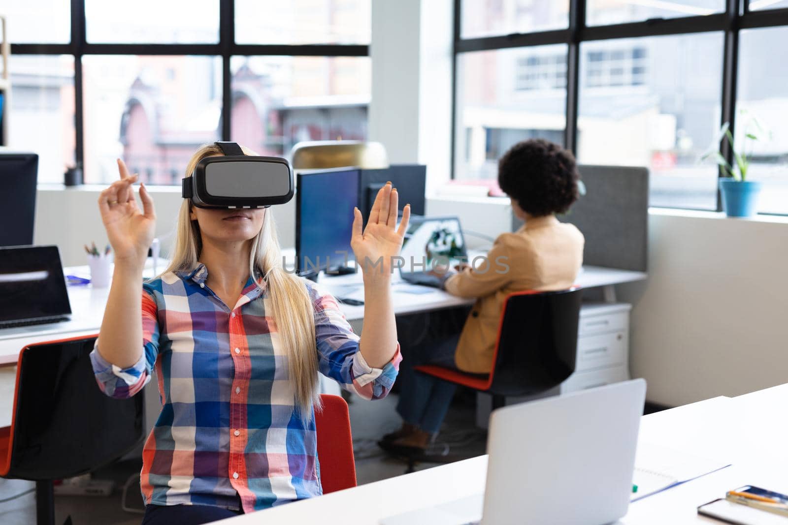 Caucasian businesswoman sitting at desk using vr goggles in creative office. with colleague in the background social distancing in workplace during covid 19 pandemic
