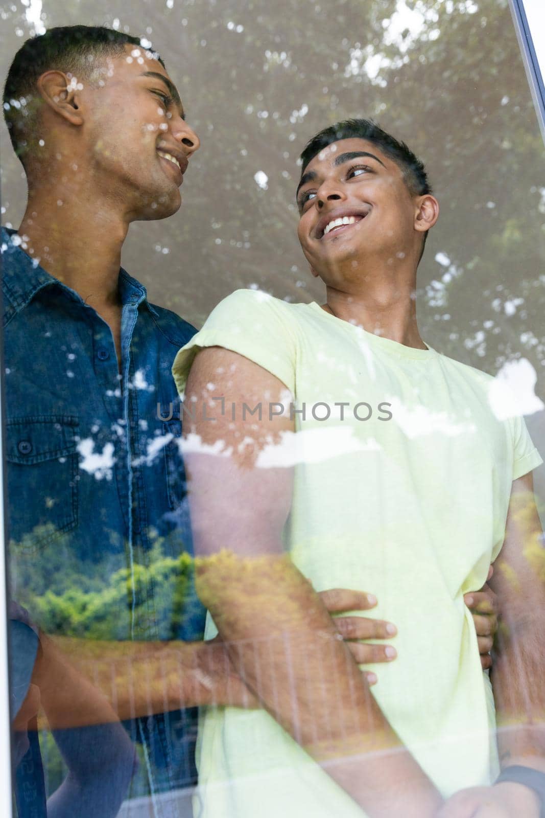 Diverse gay male couple standing by window smiling and embracing by Wavebreakmedia