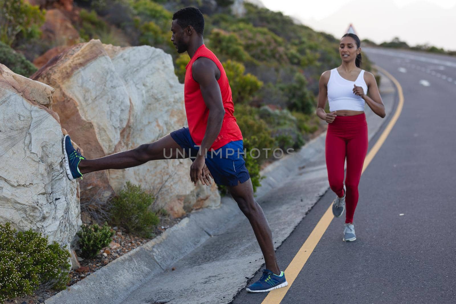 Fit african american man in sportswear stretching while woman is running on a coastal road. healthy lifestyle, exercising in nature.