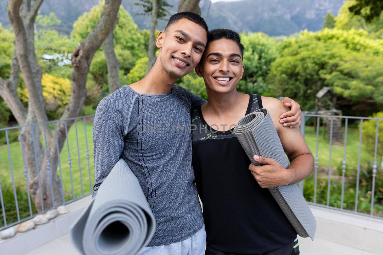 Portrait of diverse gay male couple holding yoga mats and smiling on balcony by Wavebreakmedia