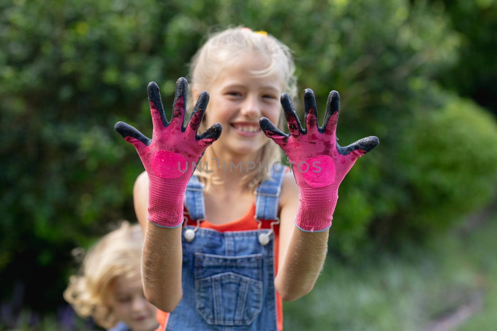 Smiling caucasian girl in garden wearing dirty pink gardening gloves, with brother in background by Wavebreakmedia