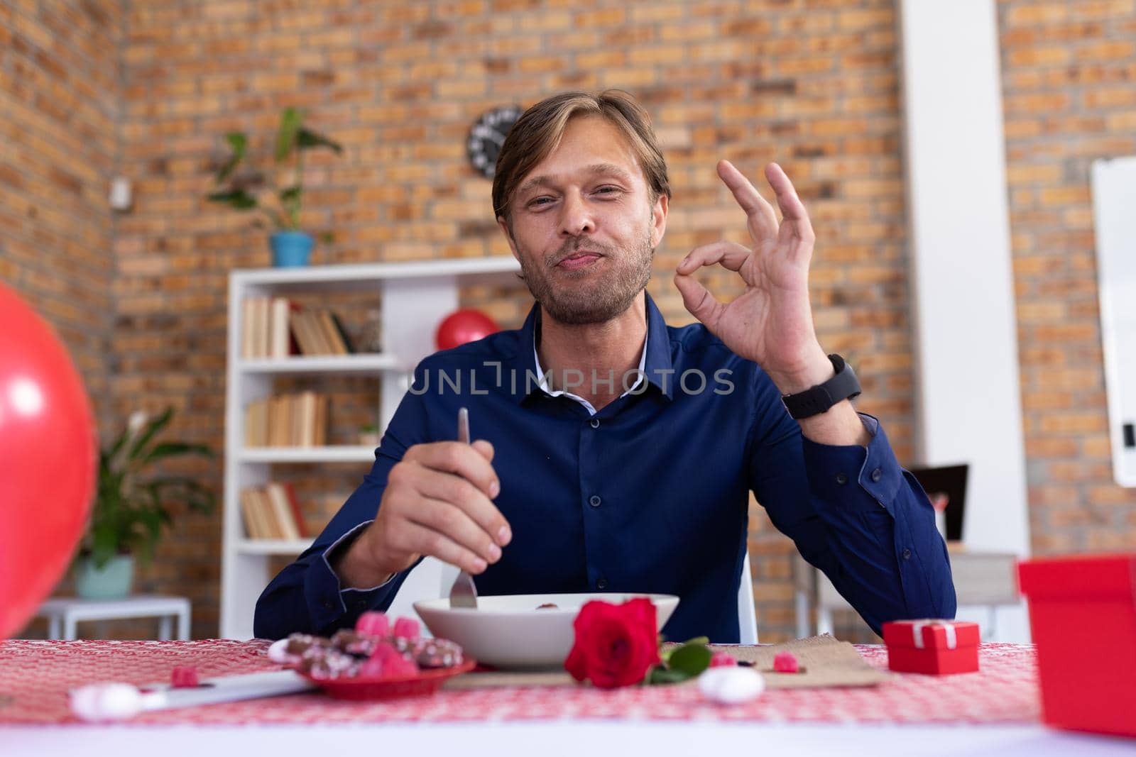 Smiling caucasian man making video call sitting at table eating making a-ok gesture. online communication during quarantine lockdown