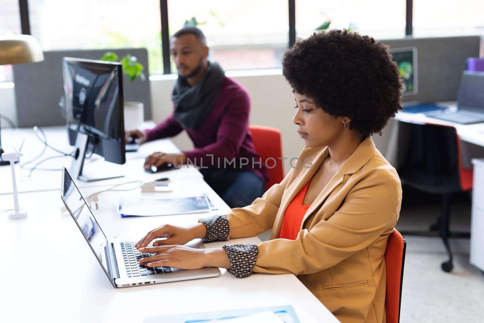 Mixed race businesswoman working in creative office. woman sitting at desk and using laptop computer, with colleague in the background. social distancing in workplace during covid 19 pandemic.