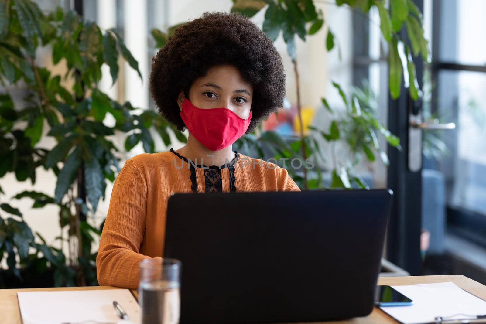 Mixed race businesswoman wearing face mask in creative office. portrait of a woman looking at camera and using laptop. social distancing protection hygiene in workplace during covid 19 pandemic.