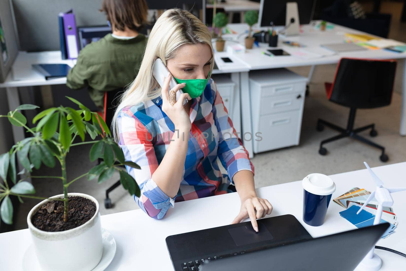 Caucasian businesswoman wearing face mask in creative office. woman sitting at desk, using laptop and talking on smartphone. social distancing protection hygiene in workplace during covid 19 pandemic.