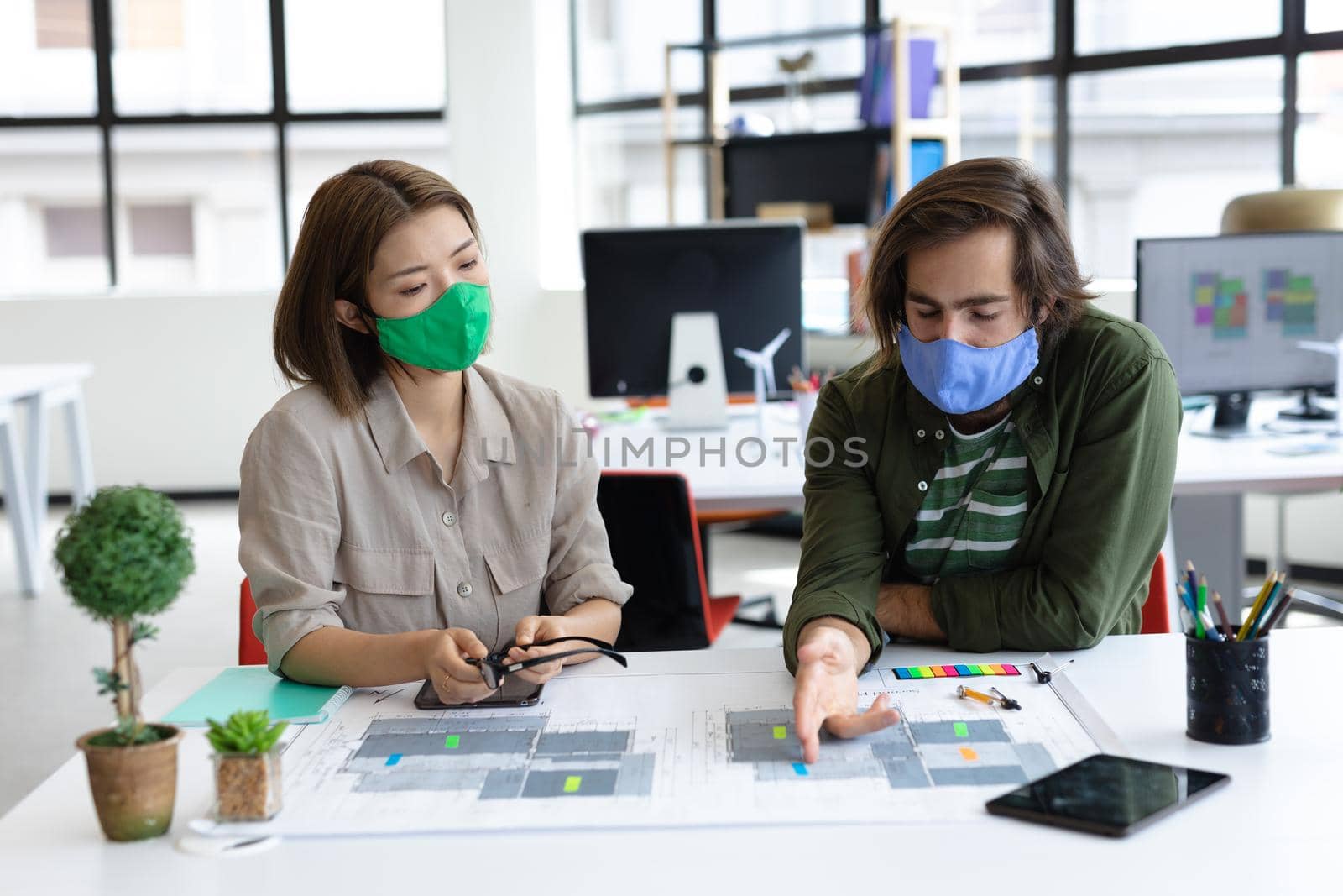 Diverse male and female colleague wearing face masks looking at blueprints and discussing by Wavebreakmedia