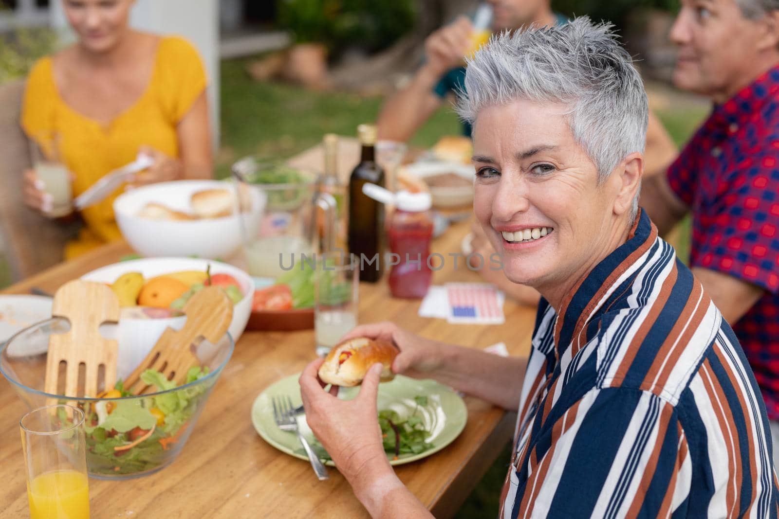 Portrait of smiling caucasian senior woman holding hotdog at table with family having meal in garden by Wavebreakmedia