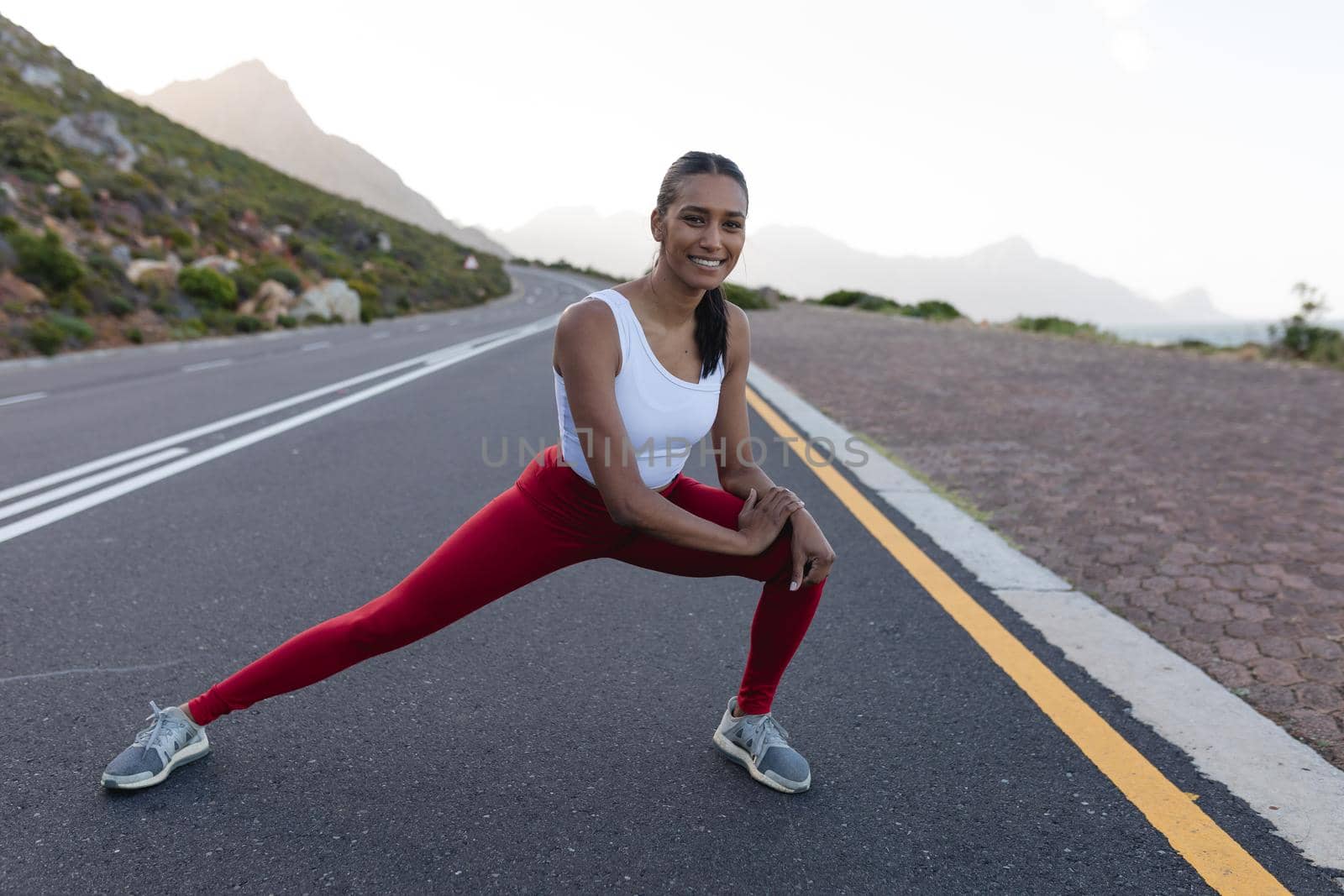 Fit african american woman in sportswear stretching on a coastal road. looking at the camera and smiling. healthy lifestyle, exercising in nature.