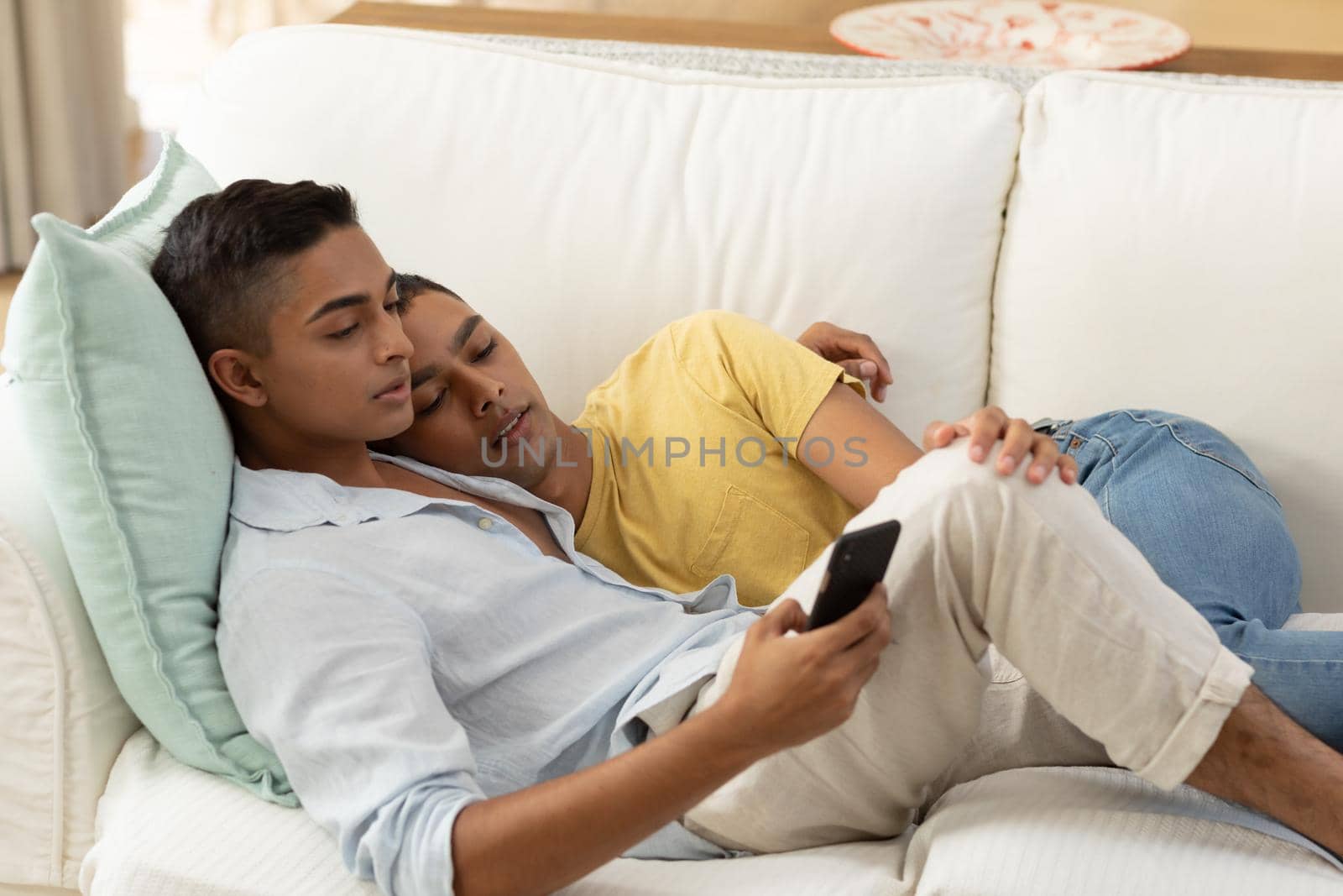 Diverse gay male couple sitting on sofa using smartphone and embracing by Wavebreakmedia