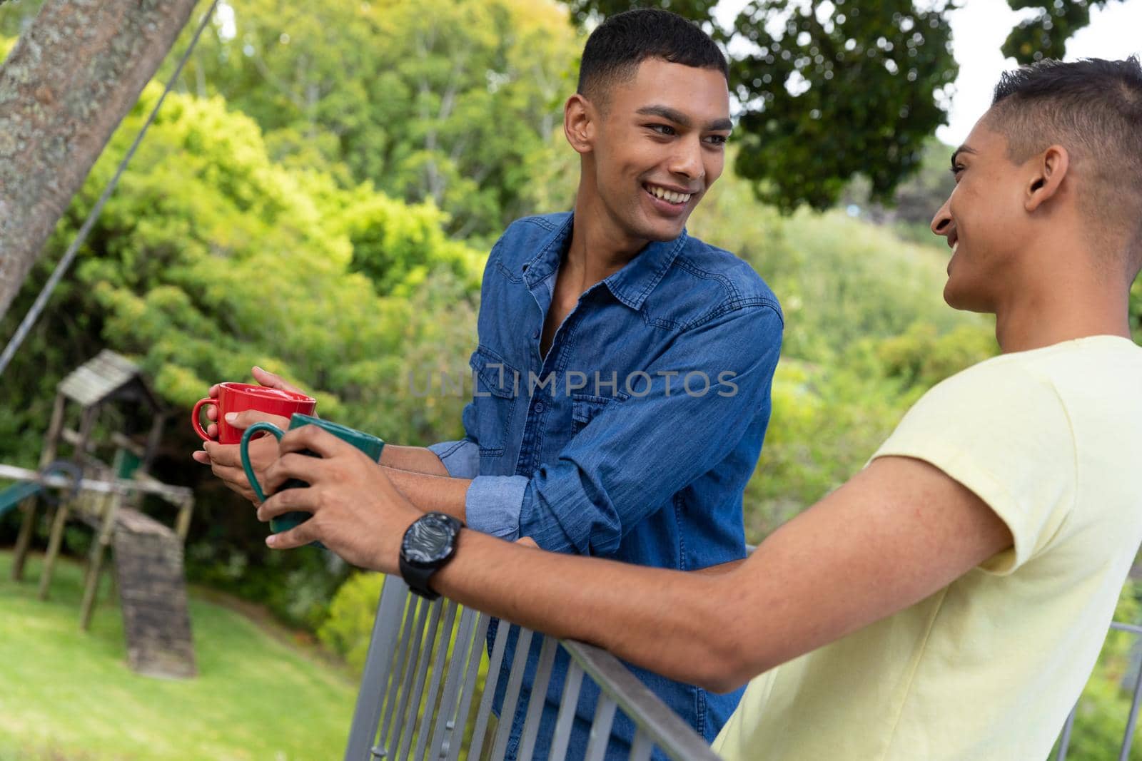 Diverse gay male couple drinking coffee and smiling on balcony. staying at home in isolation during quarantine lockdown.