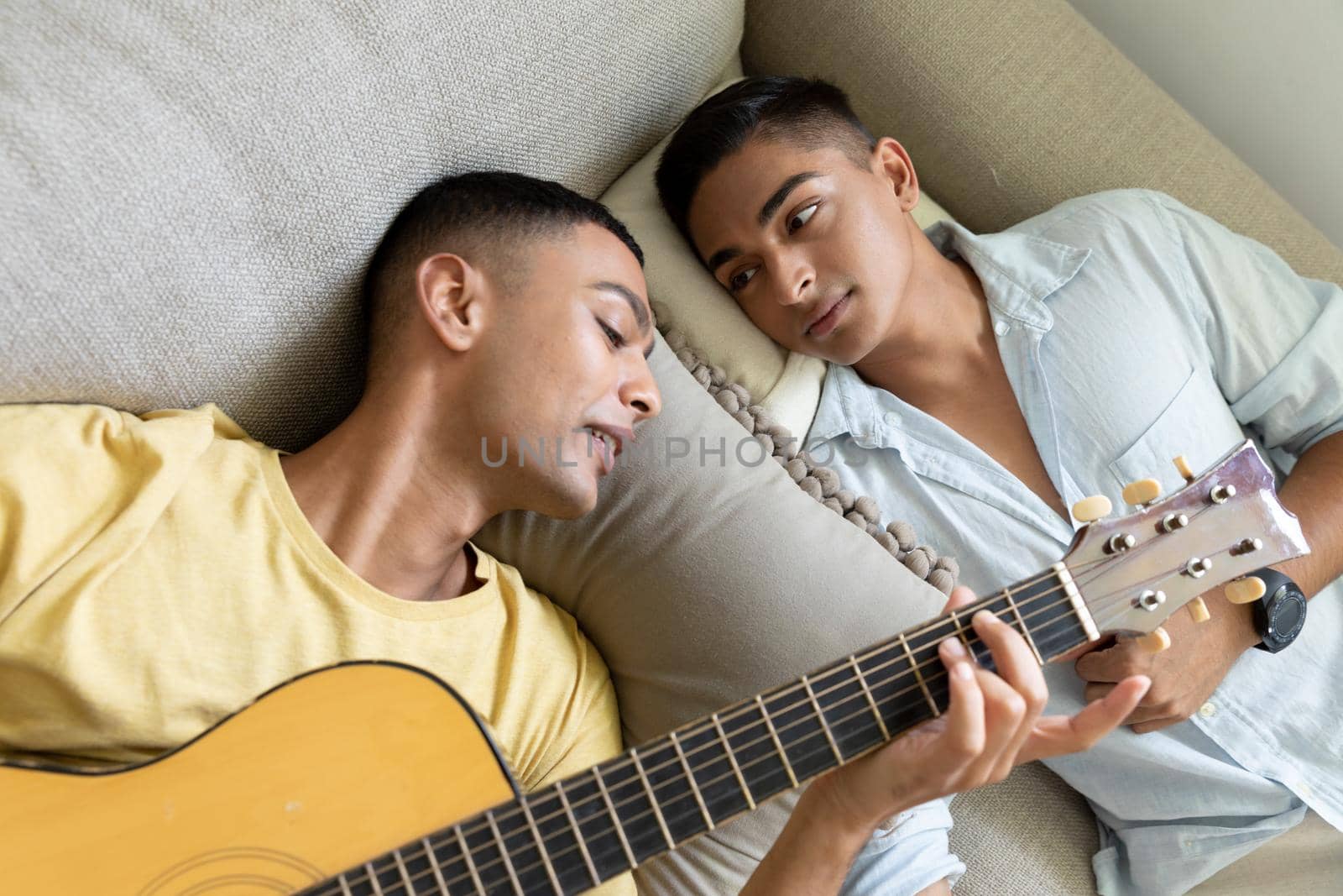 Diverse gay male couple lying on sofa playing guitar. staying at home in isolation during quarantine lockdown.