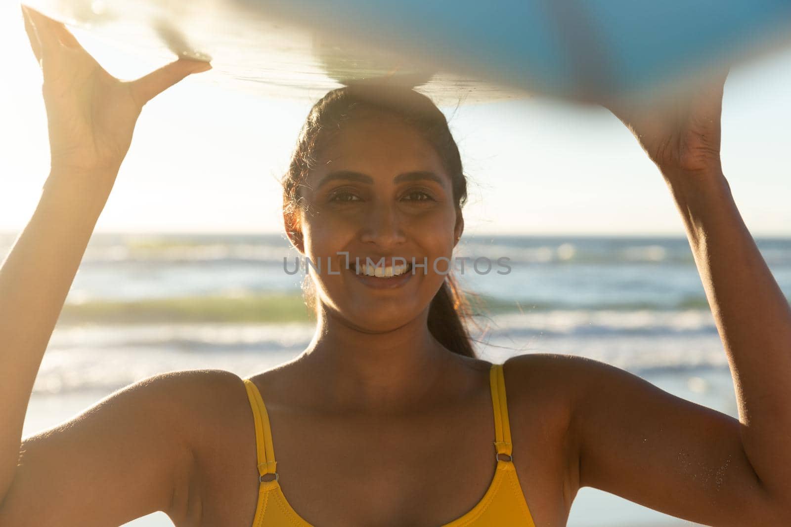 Happy mixed race woman on the beach carrying surfboard on head. healthy outdoor leisure time by the sea.