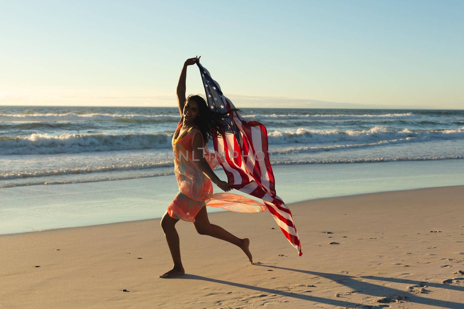 Mixed race patriotic woman running with american flag on the beach. healthy outdoor leisure time by the sea.