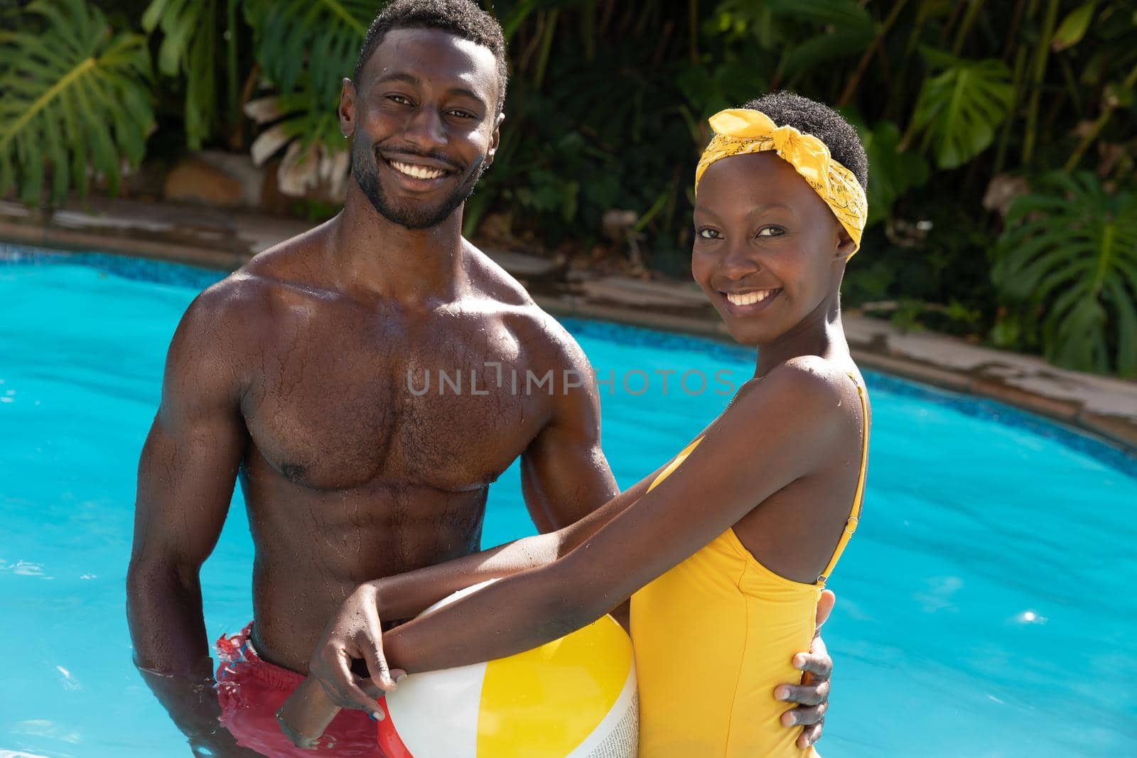 Portrait of african american couple in swimming pool holding beach ball on sunny garden terrace. staying at home in isolation during quarantine lockdown.