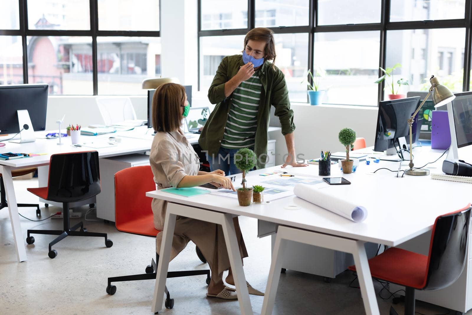 Diverse male and female colleague wearing face masks looking at blueprints and discussing by Wavebreakmedia