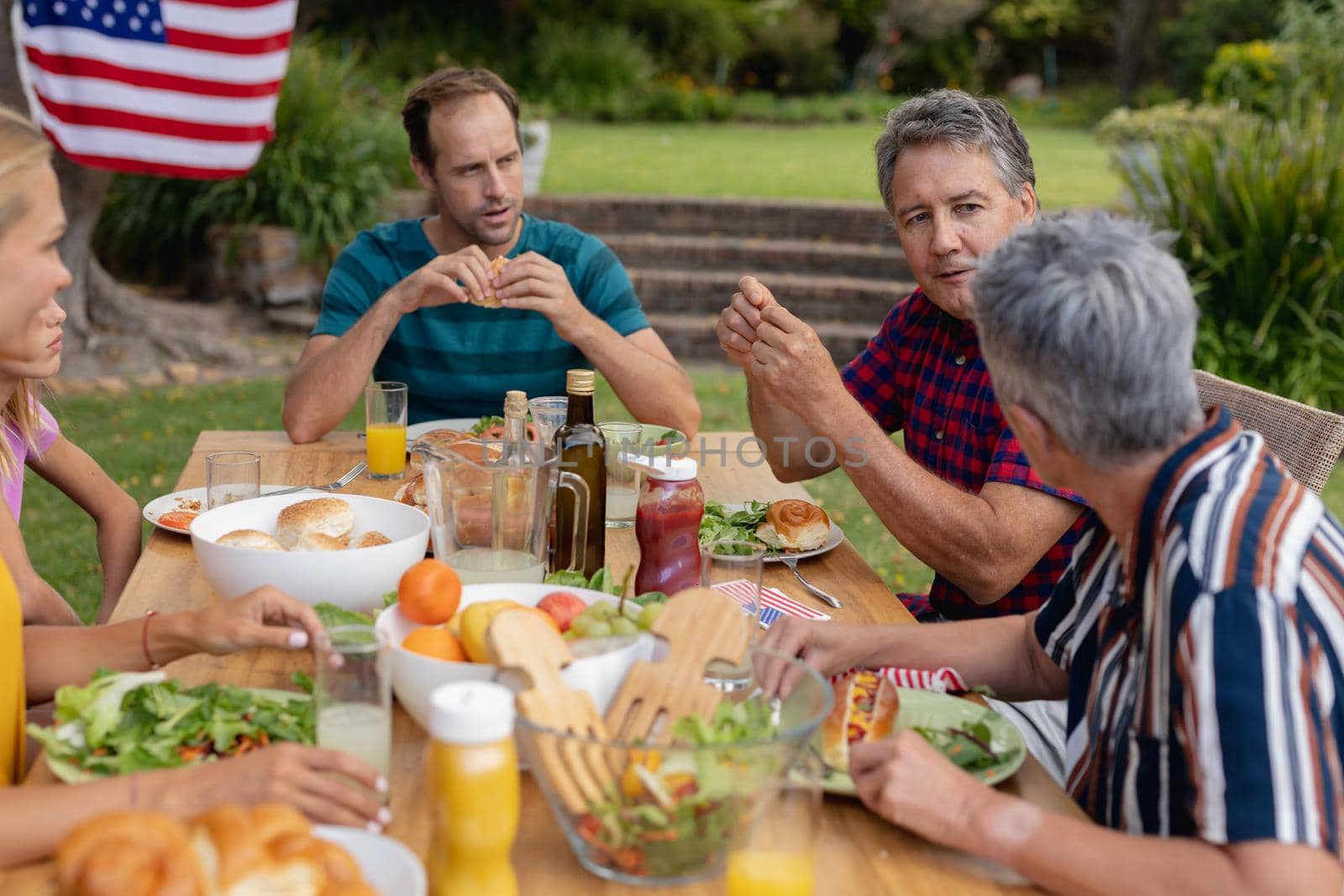Caucasian senior man sitting at table talking with family having celebration meal in garden. three generation family celebrating independence day eating outdoors together.