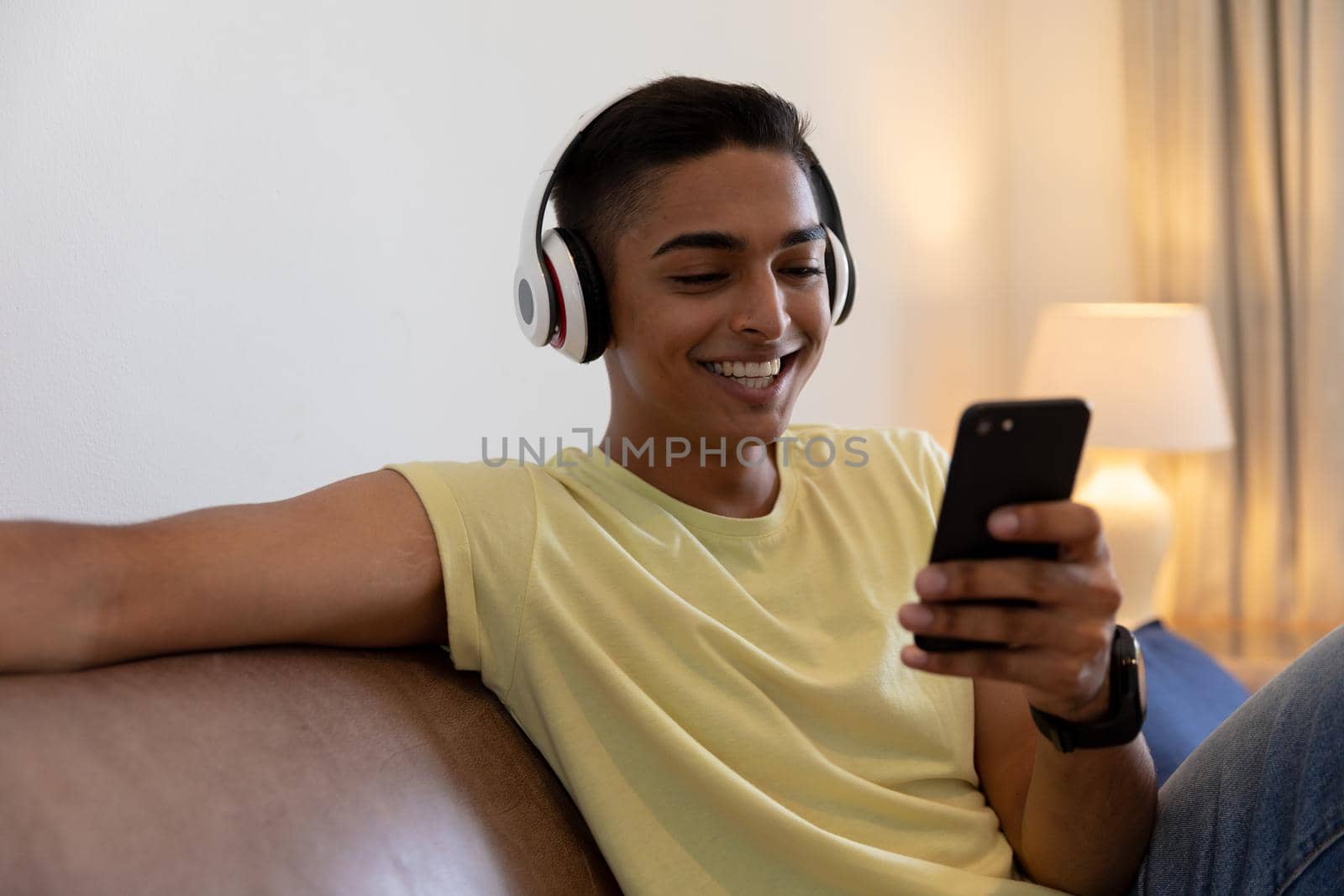 Mixed race man sitting on couch wearing headphones smiling and using smartphone. staying at home in isolation during quarantine lockdown.