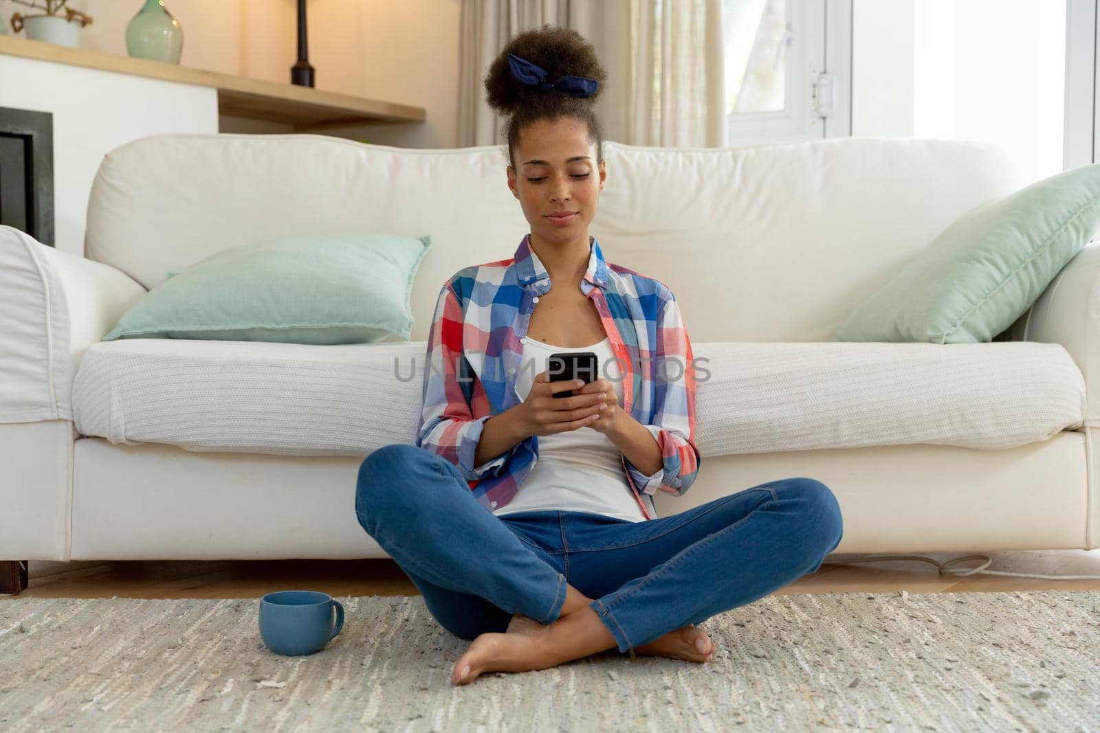 Mixed race woman sitting on the floor using smartphone and drinking coffee at home. staying at home in isolation during quarantine lockdown.