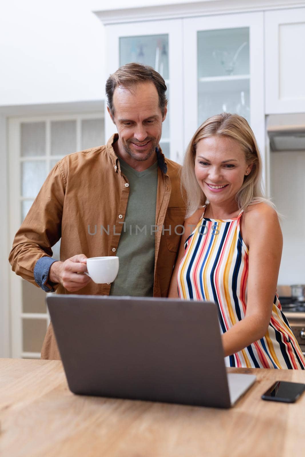 Smiling caucasian couple looking at laptop together and drinking coffee in kitchen. staying at home in isolation during quarantine lockdown.