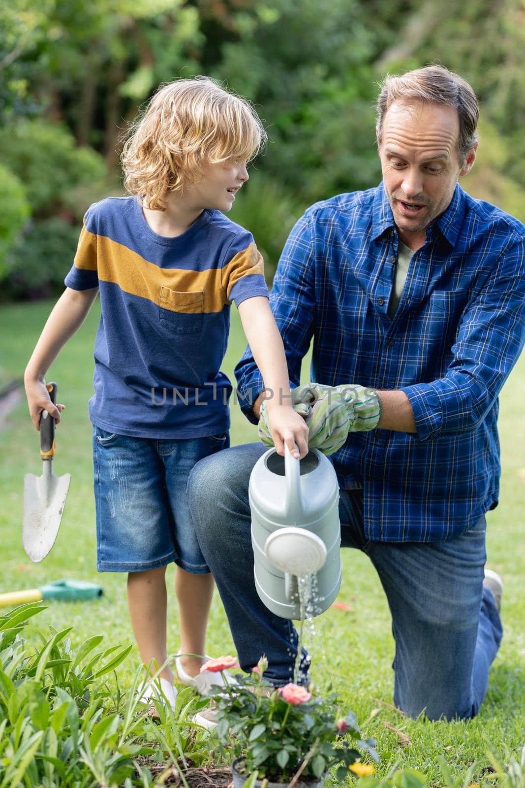 Happy caucasian father in garden with son, watering plants and gardening together. staying at home in isolation during quarantine lockdown.