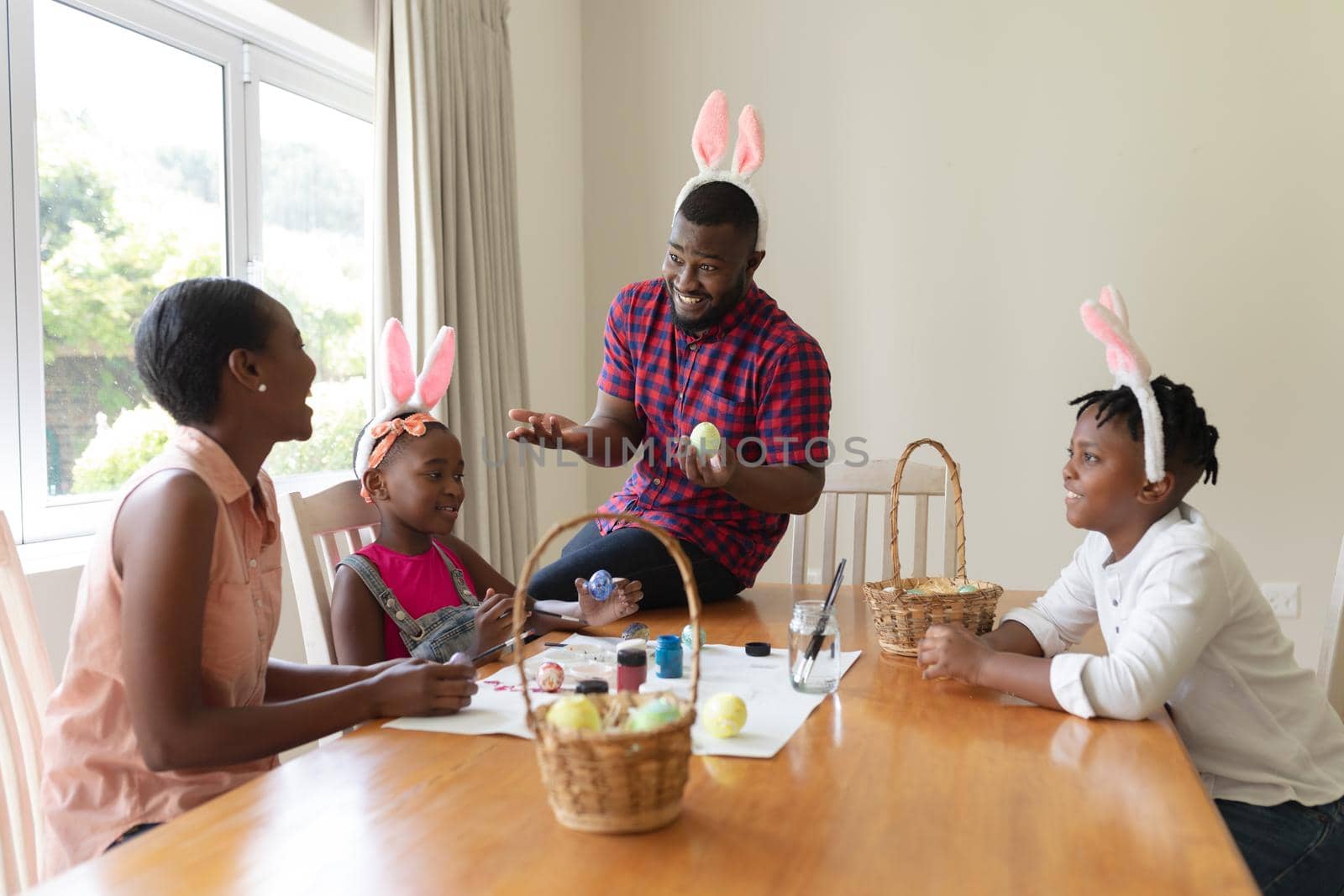 Happy african american parents with son and daughter wearing bunny ears painting easter eggs. celebrating easter at home in isolation during quarantine lockdown.