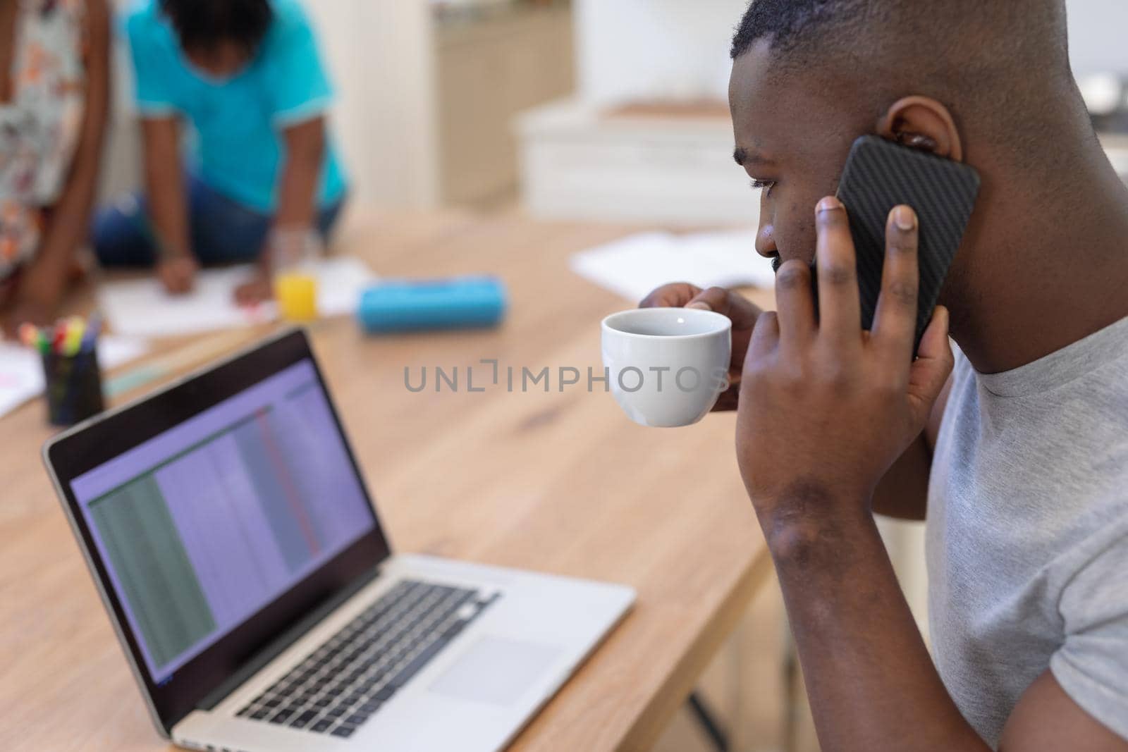 African american father working from home using laptop and smartphone, with family in background. staying at home in isolation during quarantine lockdown.