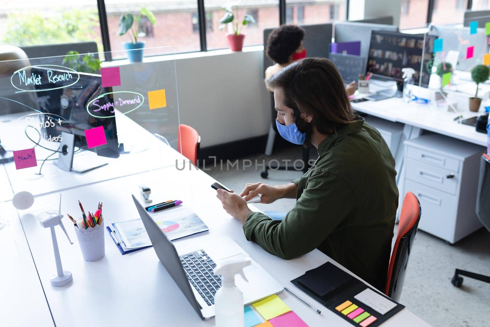 Caucasian businessman wearing face mask in creative office. man sitting at desk cleaning and disinfecting smartphone. social distancing protection hygiene in workplace during covid 19 pandemic.