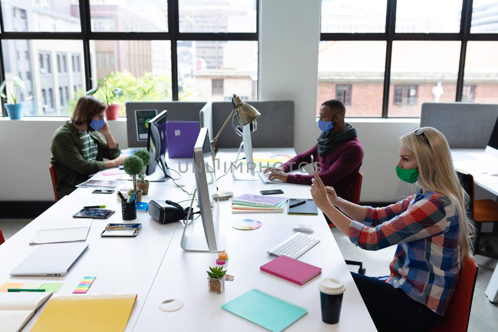 Diverse group of business people wearing face masks in creative office. group of people sitting at desks using computers. social distancing protection hygiene in workplace during covid 19 pandemic.