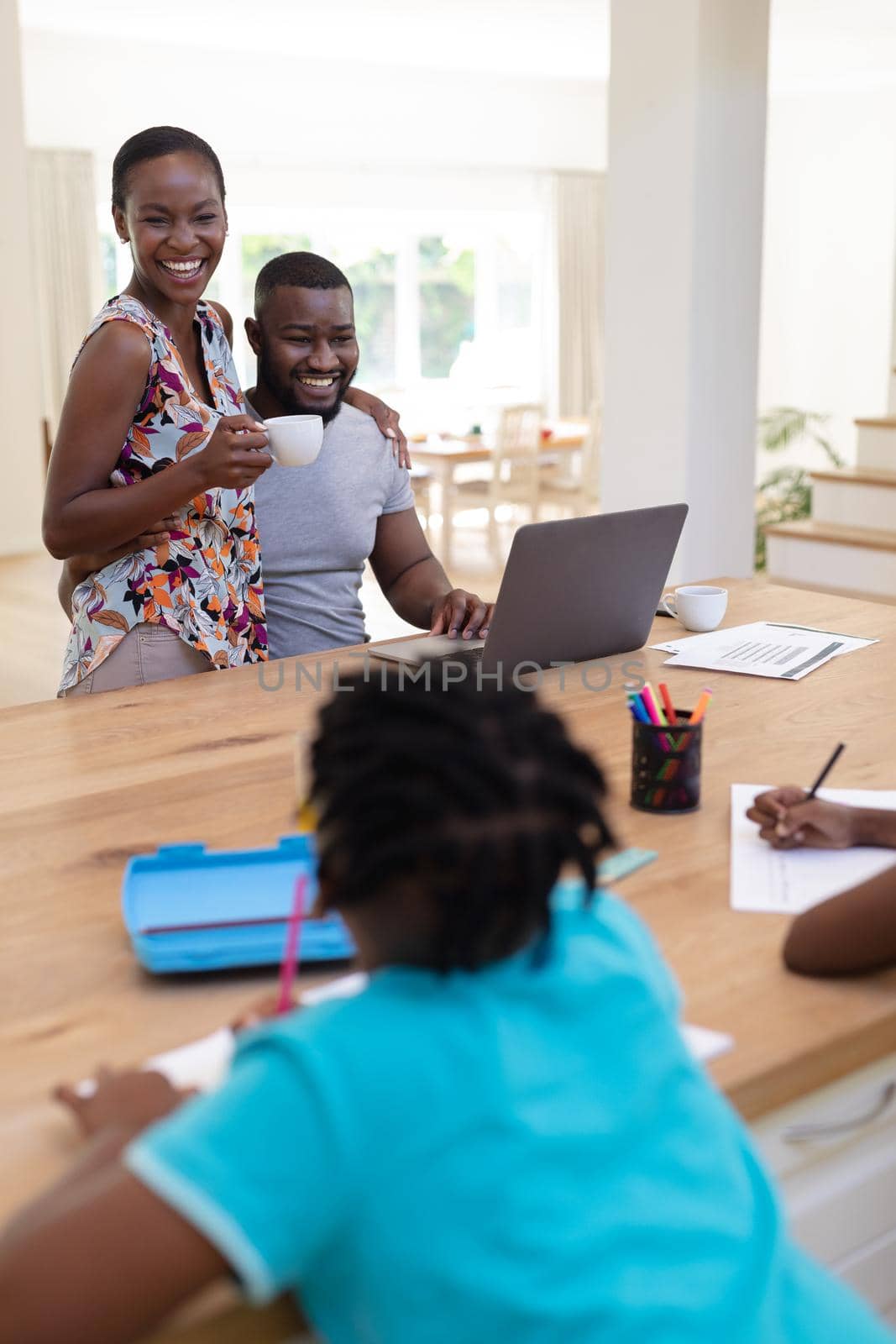 Happy african american parents using laptop embracing holding coffee with kids working at the table. staying at home in isolation during quarantine lockdown.