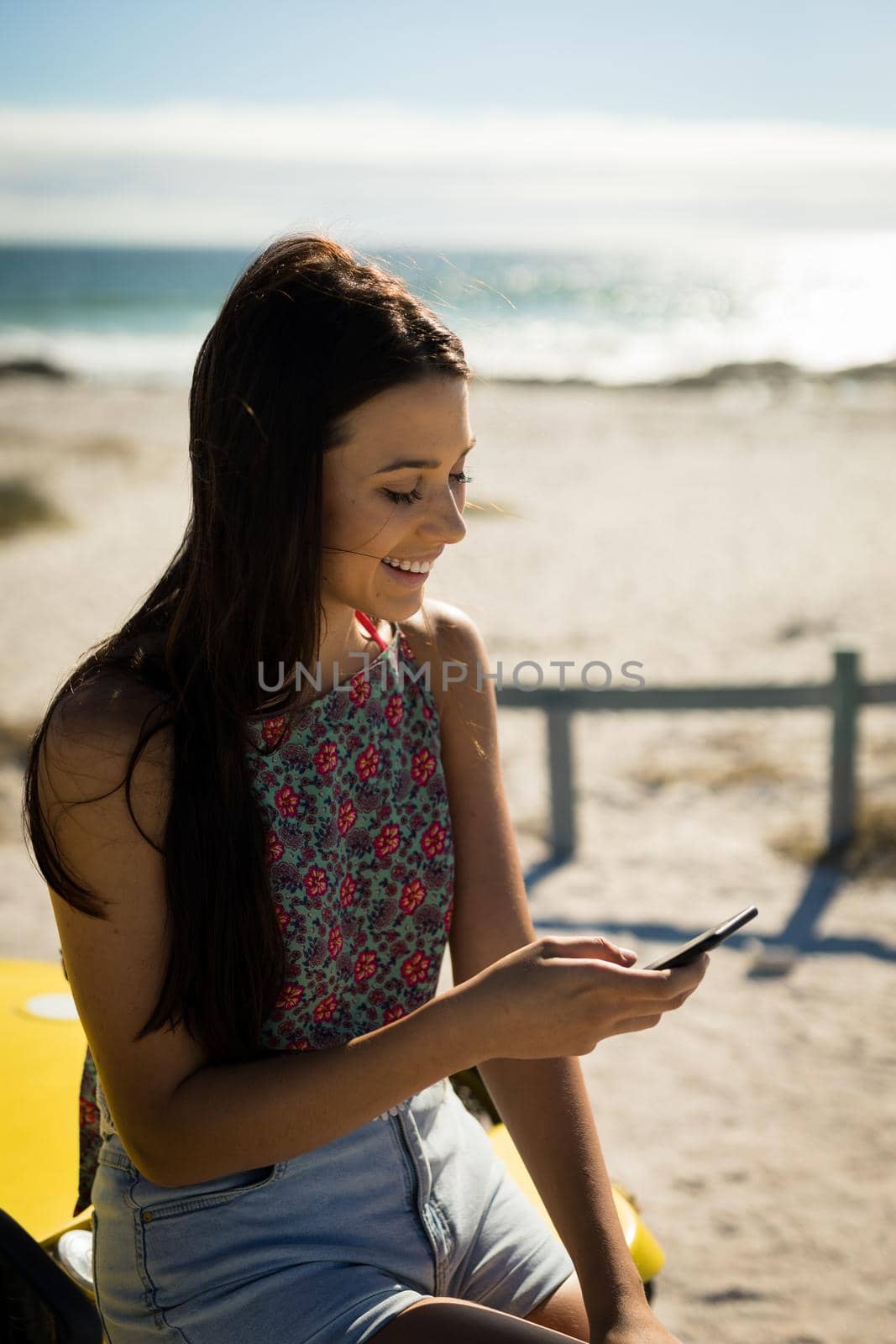 Happy caucasian woman sitting on beach buggy by the sea using smartphone. beach break on summer holiday road trip.