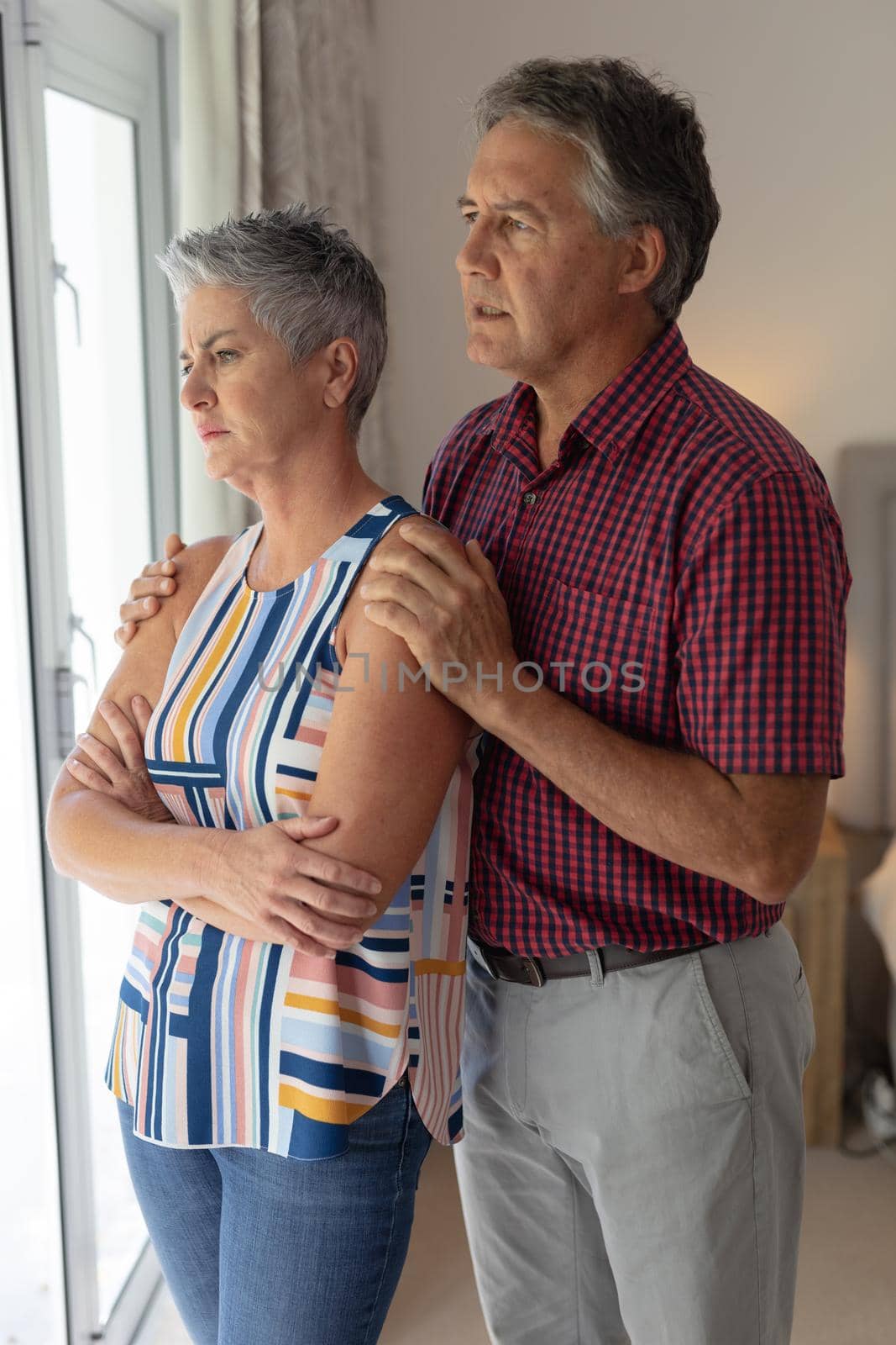 Concerned senior caucasian couple standing looking out of window embracing. staying at home in isolation during quarantine lockdown.