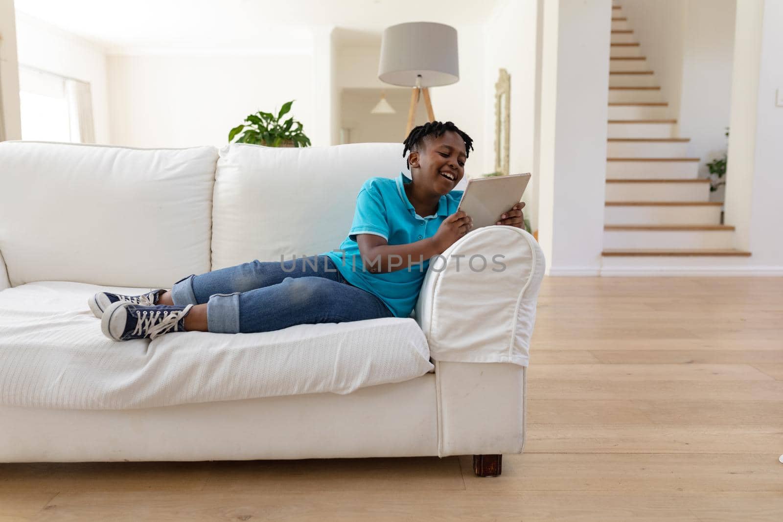 Smiling african american boy with short dreadlocks lying on couch using digital tablet. staying at home in isolation during quarantine lockdown.