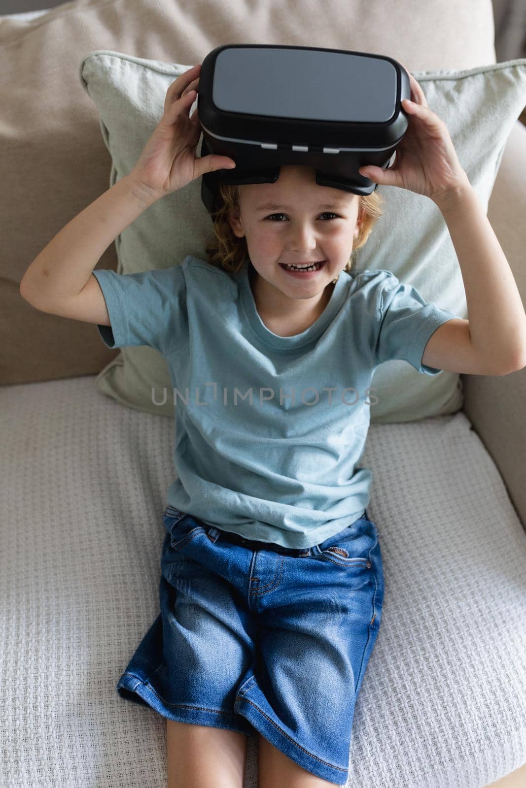 Portrait of caucasian boy sitting on couch wearing vr headset raising it and laughing. staying at home in isolation during quarantine lockdown.