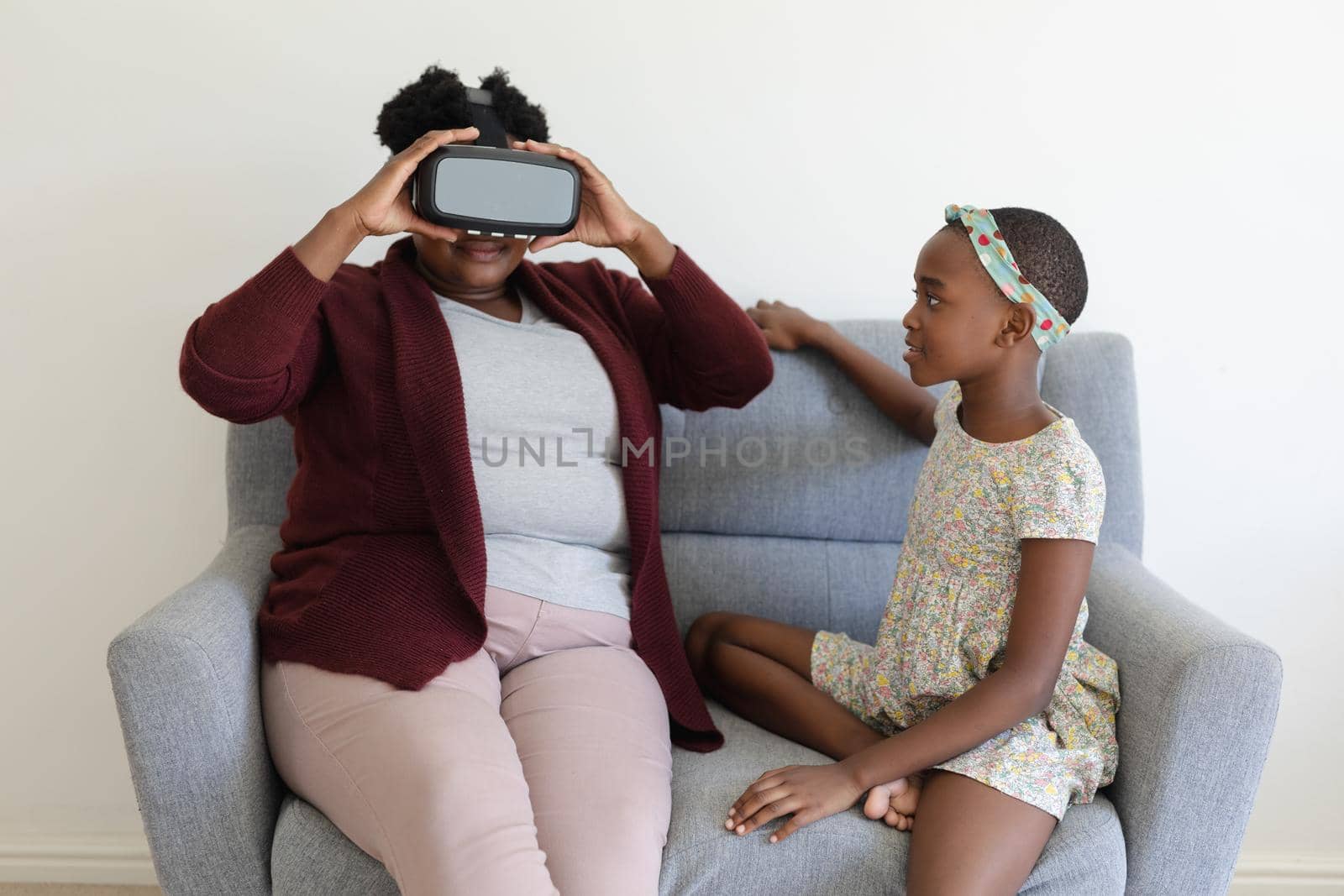 Senior african american grandmother sitting on couch wearing vr headset beside smiling granddaughter. happy family spending time together at home.