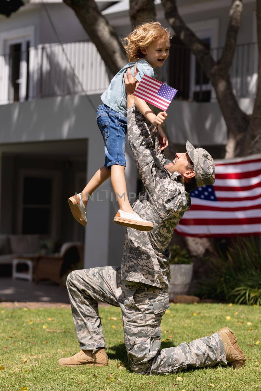 Smiling caucasian soldier father lifting happy son in garden with american flag outside house. soldier returning home to family.