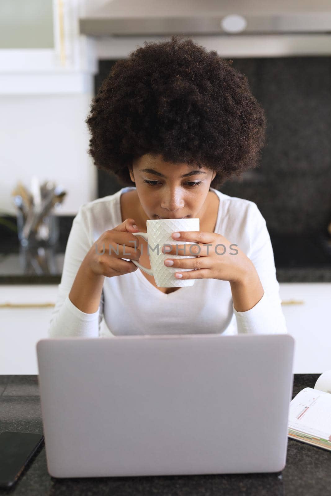 African american woman sitting in kitchen drinking coffee and using laptop by Wavebreakmedia