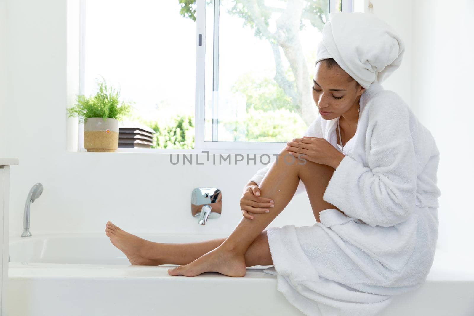 Mixed race woman wearing bathrobe and putting cream on her legs in bathroom. staying at home in isolation during quarantine lockdown.