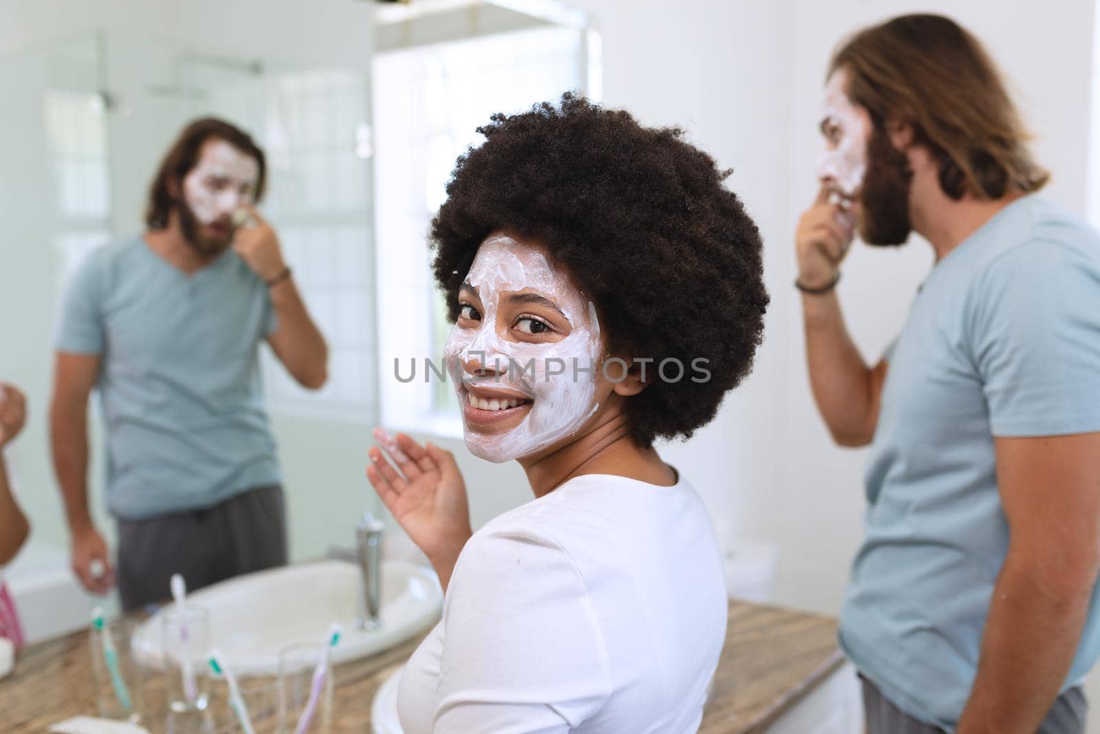 Portrait of happy diverse couple standing in bathroom wearing beauty masks. staying at home in isolation during quarantine lockdown.