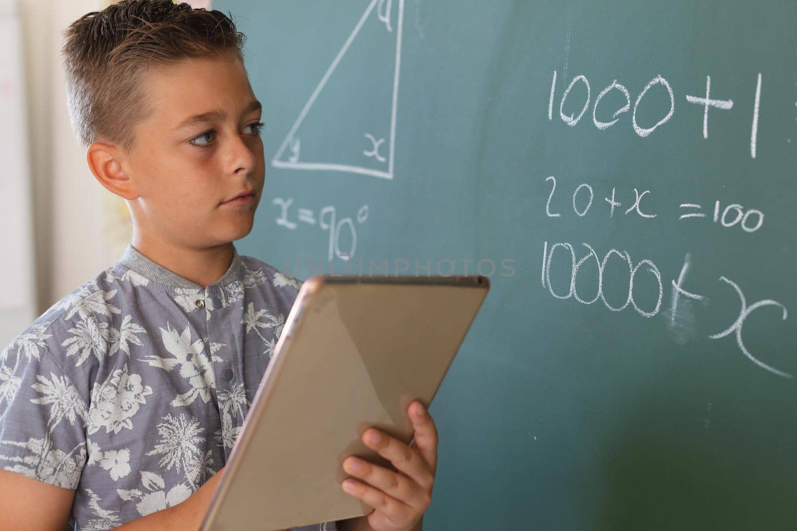 Caucasian boy standing at chalkboard in classroom and using tablet during maths lesson. childhood and education at elementary school.