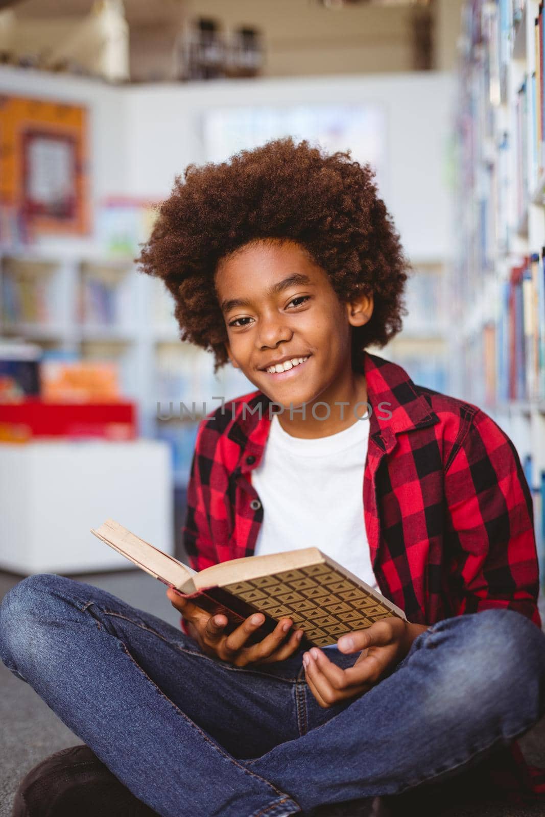 Portrait of smiling african american schoolboy reading book sitting on floor in school library by Wavebreakmedia