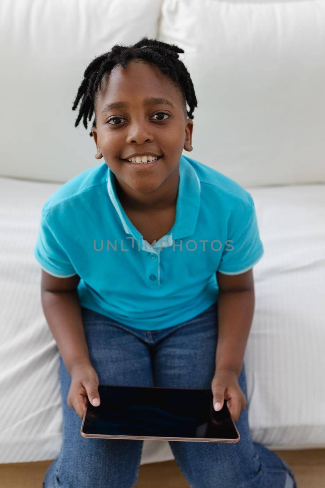Portrait of smiling african american boy with short dreadlocks sitting on couch using digital tablet by Wavebreakmedia