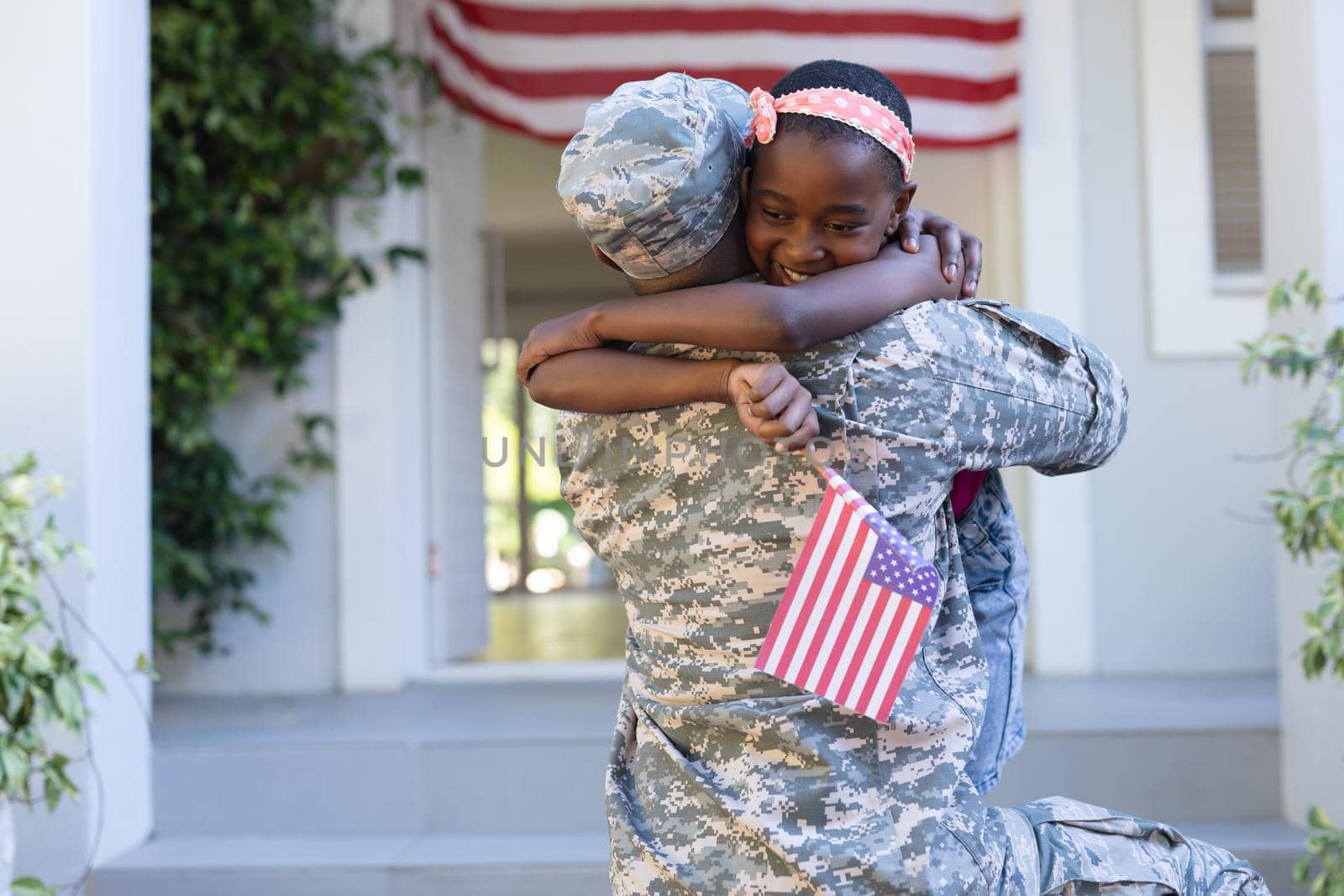 African american soldier father hugging smiling daughter holding flag in front of house by Wavebreakmedia