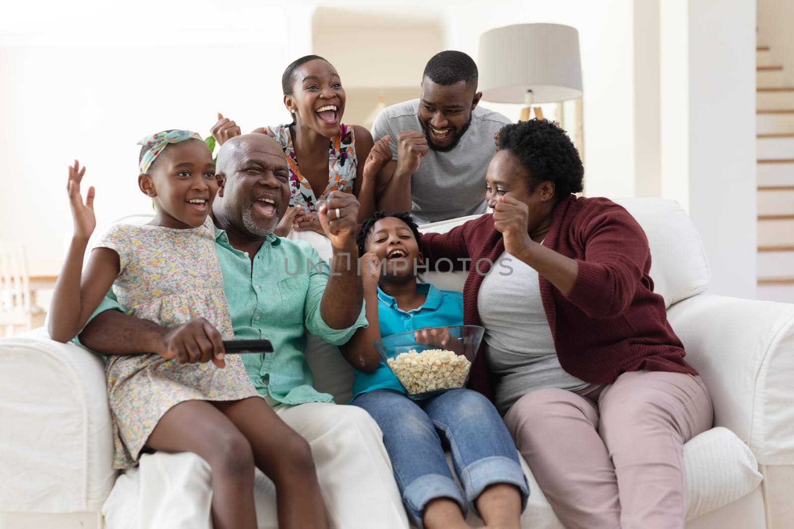 Excited african american parents, grandparents and grandchildren on couch watching tv and cheering. happy three generation family spending time together at home.