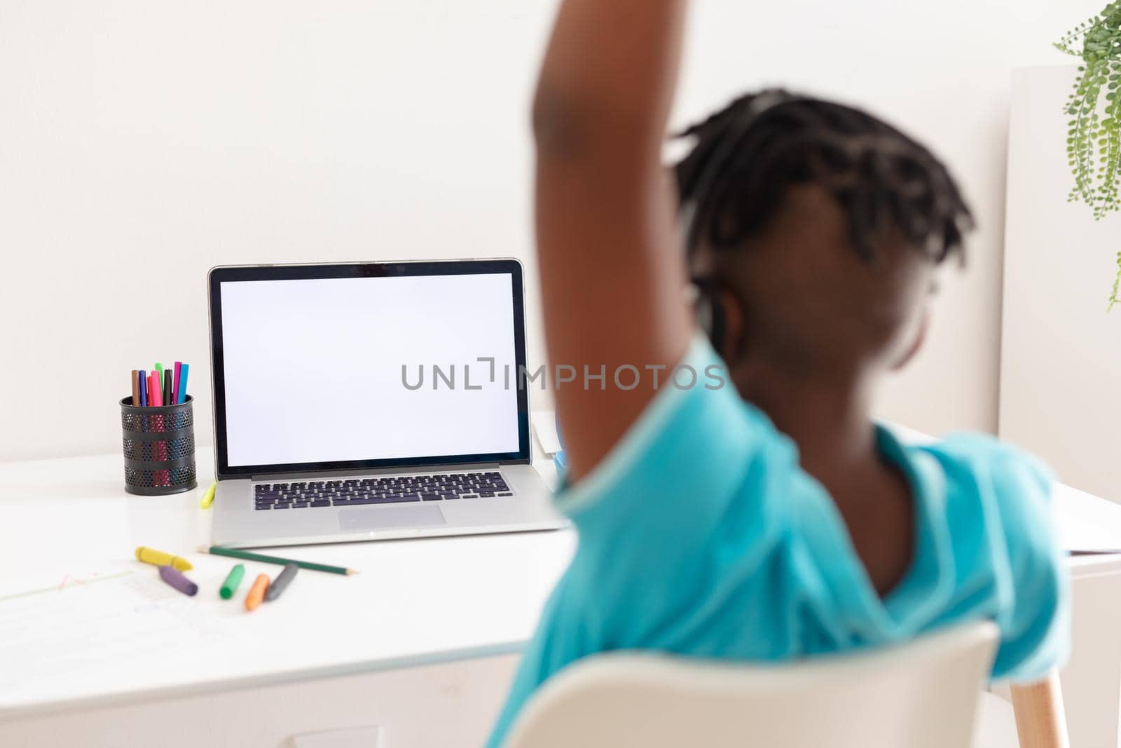 African american boy at desk using laptop with copy space for online school lesson and raising hand. staying at home in isolation during quarantine lockdown.