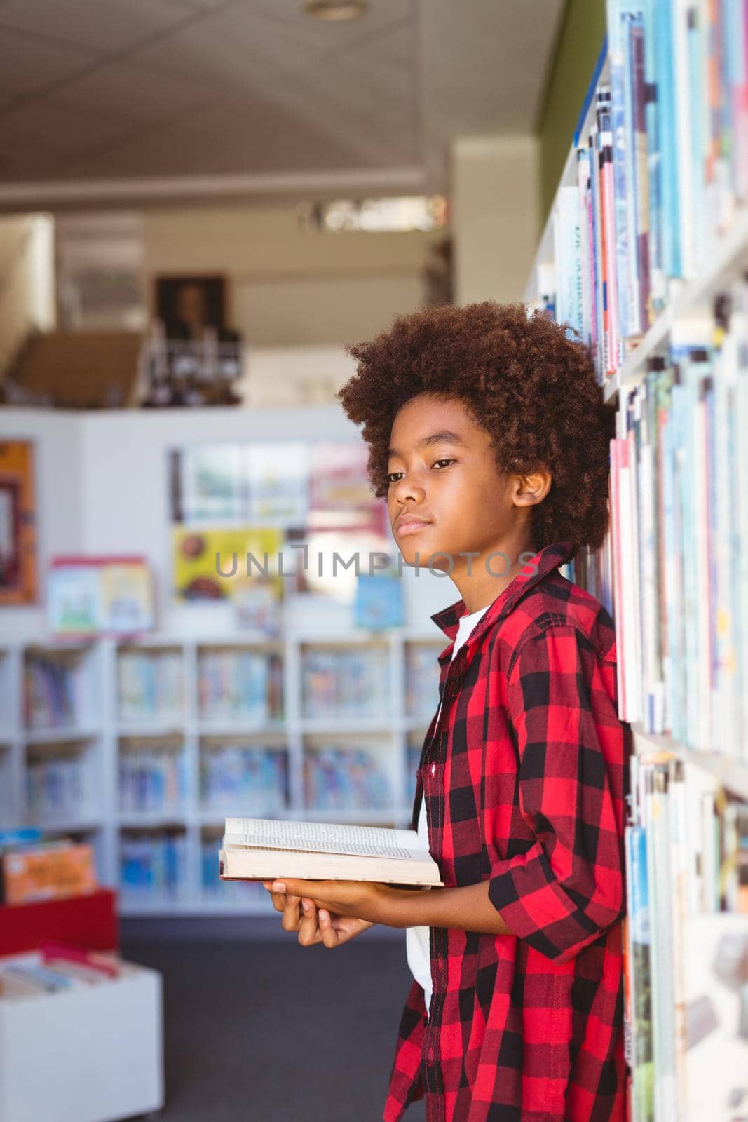 Happy african american schoolboy reading book standing in school library, looking away. childhood and education at elementary school.