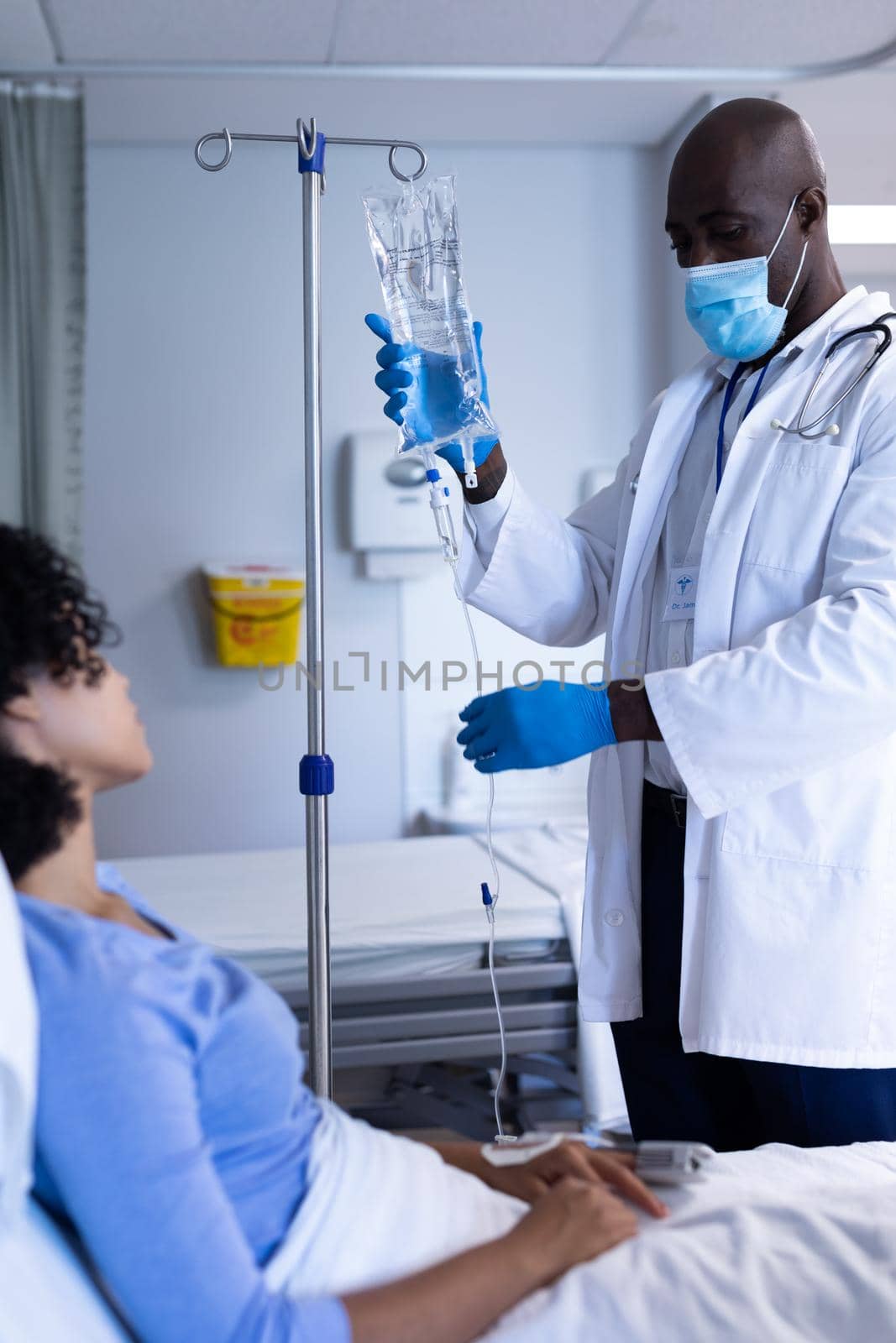 African american male doctor wearing face mask preparing iv bag for patient sitting in hospital bed. medicine, health and healthcare services during coronavirus covid 19 pandemic.